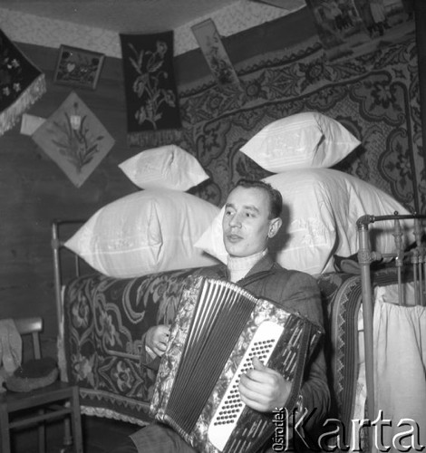 Styczeń 1958, Augustowo, pow. Bielsk-Podlaski, woj. Białystok, Polska.
Młody mężczyzna z akordeonem siedzi na tle łóżka, na którym leżą poduszki. 
Fot. Romuald Broniarek, zbiory Ośrodka KARTA