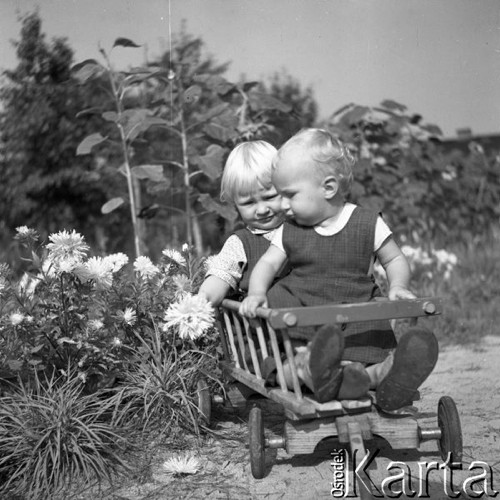 Lipiec 1955, Puławy, Polska.
Dwoje dzieci siedzi w drewnianym wózku stojącym w ogrodzie pośród kwiatów.
Fot. Romuald Broniarek, zbiory Ośrodka KARTA