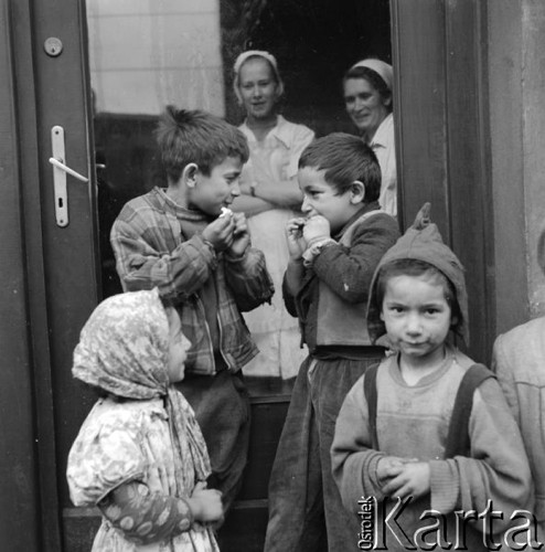Kwiecień 1958, Warszawa, Polska. 
Cygańskie dzieci jedzą bułkę przed kuchnią stołówki. Za zamkniętymi drzwiami stoją dwie kobiety w fartuchach.
Fot. Romuald Broniarek, zbiory Ośrodka KARTA