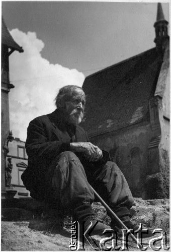 1947, Polska.
Stary mężczyzna z laską siedzący na ziemi, w tle kościół, na odwrocie podpis autora 