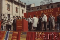 Lata 80., Włochy.
Pierwszomajowa demonstracja zorganizowana przez włoskie związki zawodowe. Manifestanci stoją na udekorowanym na czerwono podium. Widoczni są m.in. przemawiająca kobieta oraz mężczyzna z kamerą (po lewej stronie). W tle, na budynku widnieje reklama 