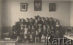 Brak daty, brak miejsca.
Zdjęcie grupowe dzieci ubranych w szkolne fartuszki i mundurki marynarskie. Po lewej stronie siedzi ksiądz, po prawej - opiekunka dzieci. Na ścianie zawieszone zostało Godło Państwowe oraz portret Marszałka Józefa Piłsudskiego (po lewej stronie).
Fot. Józef Tarań, zbiory Ośrodka KARTA, kolekcję zdjęć przekazała Lucyna Kumiszczo