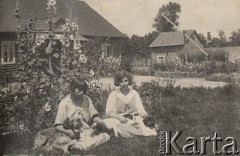 1924-1932, Kosów Poleski, woj. poleskie, Polska.
Dwie dziewczyny z psami siedzące na trawie przed domem na tle kwiatów malwy, z prawej studnia z żurawiem.
Fot. Feliks Drozdowski, zbiory Ośrodka KARTA, Pogotowie Archiwalne [PAF_050], udostępnił Krzysztof Drozdowski