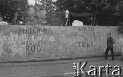 Sierpień 1980, Gdańsk, Polska.
Strajk okupacyjny w Stoczni Gdańskiej im. Lenina. Robotnicy siedzą na murze otaczającym stocznię. Na murze hasła: 