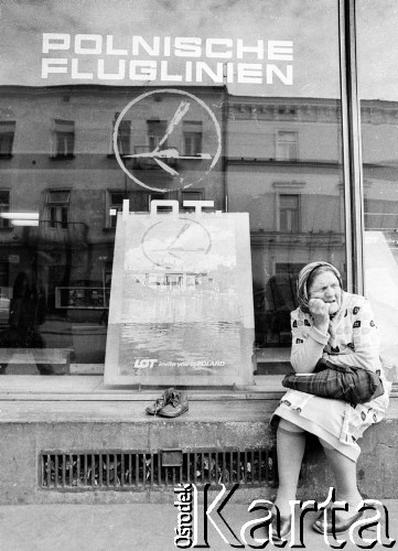 1982, Kraków, Polska.
Kobieta w chustce na głowie siedzi przed siedzibą LOT-u. Obok niej reklama i dziecięce buty.
Fot. Jerzy Szot, zbiory Ośrodka KARTA