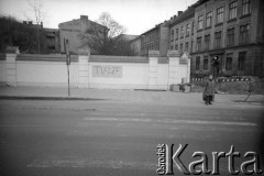 1989, Kraków, Polska.
Kobieta czeka przed przejściem dla pieszych przy murowanym ogrodzeniu, na którym widnieje napis 