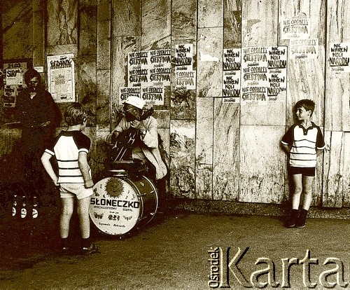 lata 80., Polska.
Chłopcy przysłuchują się mężczyźnie grającemu na gitarze. Na ścianie plakaty 