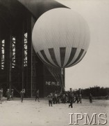 Lata 30., Legionowo, Polska.
Balon kulisty przed hangarem. Miejscowy 2. Batalion Balonowy dysponował obszernym portem balonowym. Jego najważniejszym elementem był drewniany hangar o konstrukcji szkieletowej, wzniesiony na początku lat 20. Kierownikiem nadzorującym jego budowę był inż. Stolarczyk. Hangar miał długość ok. 80 m, szerokość ok. 40 m i wysokość ok. 25 m. Mógł jednorazowo pomieścić 6-7 napełnionych balonów kulistych 750 m³. Przed hangarem rozciągał się obszerny plac startowy (widoczny częściowo na zdjęciu). Było to główne miejsce ćwiczeń legionowskich baloniarzy. Przy hangarze wybudowano także magazyny wytwórni wodoru oraz powłok balonowych. Poza funkcjami służbowymi hangar pełnił także ważną rolę podczas świąt batalionowych. Tu odbywały się nabożeństwa polowe przy ołtarzu wykonanym z balonów zaporowych częściowo napełnionych wodorem, a w czasie święta jednostki - 15 września dowódca wręczał tu nominacje na kolejny stopień wojskowy, odznaczenia i listy gratulacyjne. Współcześnie na miejscu hangaru stoją bloki przy ul. Zegrzyńskiej 11 i 19 na osiedlu Piaski w Legionowie.
Album balonowowy - fotografie po dr Niedźwierskim.
Fot. NN, Instytut Polski im. Gen. Sikorskiego w Londynie
