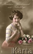 Pocz. XX wieku, Francja.
Portret kobiety w kolorowej sukni, z naszyjnikiem na szyi, stojącej przy stroiku z bukietem kwiatów.  Napis w języku niemieckiem: 