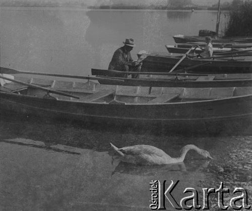 1942-1943, Moosseedorf, Szwajcaria.
Mężczyzna czyta gazetę siedząc na łódce przy brzegu. W łódce obok siedzi dziewczynka i trzyma wiosła. Do łódek podpływa łabędź. 
Fot. Jerzy Konrad Maciejewski, zbiory Ośrodka KARTA
 
