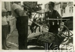 Lata 30., prawdopodobnie Bułgaria.
Dwaj mężczyźni trzymają drewniany pałąk, na którym jest zaczepiony na łańcuchach ciężki worek. Z tyłu za nimi stoi wóz zaprzęgnięty w osła. 
Fot. Jerzy Konrad Maciejewski, zbiory Ośrodka KARTA