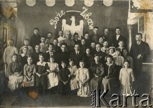 Ok. 1920, brak miejsca.
Jan Piekutowski (w 2 rzędzie, 5 z lewej) siedzi wśród dzieci i młodzieży oraz ich opiekunów. W tle dekoracja z wizerunkiem orła białego w koronie i napisem 