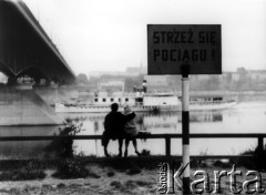 Wrzesień 1962, Warszawa, Polska.
Obejmująca się para siedzi nad brzegiem Wisły pod mostem. Na pierwszym planie tabliczka z napisem 
